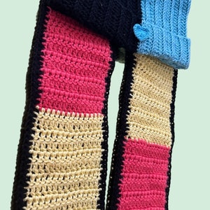 May include: A crocheted scarf with black, pink, beige, and blue sections. The scarf is made of different colored squares sewn together. The scarf is folded in half and the ends are hanging down.