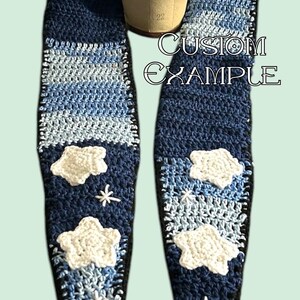 May include: A blue crocheted hat and scarf set with white crocheted clouds and stars. The scarf has a gradient of blue colors. The hat has a white flower on top. The set says "CUSTOM EXAMPLE".