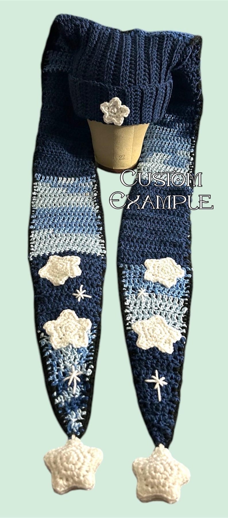 May include: A blue crocheted hat and scarf set with white crocheted clouds and stars. The scarf has a gradient of blue colors. The hat has a white flower on top. The set says "CUSTOM EXAMPLE".