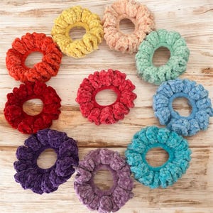 May include: A collection of colorful, plush scrunchies in various shades, including red, yellow, pink, green, blue, purple, and teal. These soft, fabric hair accessories are arranged on a light wood-grain surface.