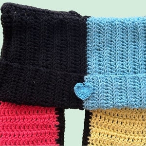 May include: A crocheted scarf with black, pink, yellow, and blue stripes. The scarf has a heart-shaped embellishment on the blue section.