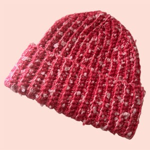 May include: A red and white knit beanie hat. The hat is made of a ribbed knit fabric with white flecks throughout. The brim is folded up, and the hat is angled slightly.
