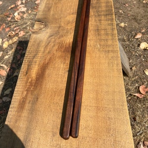 May include: A pair of dark brown wooden chopsticks with a natural wood grain pattern. The chopsticks are resting on a wooden surface.