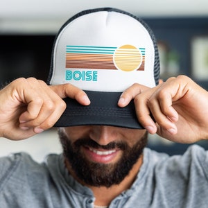May include: A white and black trucker hat with a retro-style graphic design featuring a sun and the word "BOISE" in teal blue. The hat has a black mesh back.