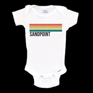 May include: White baby onesie with a retro-style rainbow stripe and the text "SANDPOINT" in black.