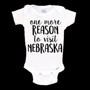 May include: White baby onesie with black text that reads "one more reason to visit NEBRASKA".