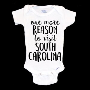 May include: White baby onesie with the text "one more REASON to visit SOUTH CAROLINA" in black. The onesie has short sleeves and snaps at the bottom. The brand name "Gerber" is visible on the tag.