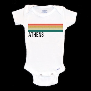 May include: White baby grow with a horizontal stripe design in shades of red, yellow, and green. The text "ATHENS" is printed in black below the stripes.