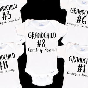 May include: Five white baby onesies with black text. The onesies announce the arrival of a grandchild with a number and month. The text reads: "Grandchild #3 Coming in November!" "Grandchild #11 Coming in July!" "Grandchild #8 Coming Soon!" "Grandchild #6 Coming in December!" "Grandchild #1 Coming in January!"