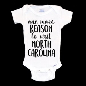 May include: White baby onesie with black text that reads "one more reason to visit North Carolina".