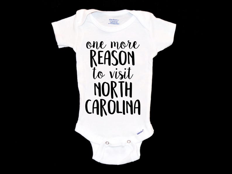 May include: White baby onesie with black text that reads "one more reason to visit North Carolina".