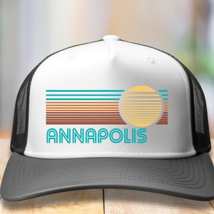 May include: White trucker hat with a black mesh back. The front of the hat features a retro-style graphic with a sun and the word "ANNAPOLIS" in blue and teal.