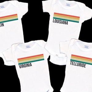 May include: Four white baby onesies with a colorful striped design and the words "Austin", "Louisiana", "Virginia", and "Telluride" printed on them.
