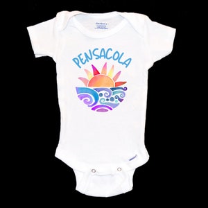 Peut inclure: Combinaison blanche pour bébé avec un motif bleu et orange d'un soleil couchant sur des vagues. Le texte "PENSACOLA" est écrit en bleu au-dessus du motif.