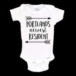 May include: White baby onesie with black text that reads "Portland's newest resident" with arrows above and below the text.