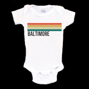 May include: White baby onesie with a retro-style striped design in red, orange, yellow, and green. The text "BALTIMORE" is printed in black below the stripes.