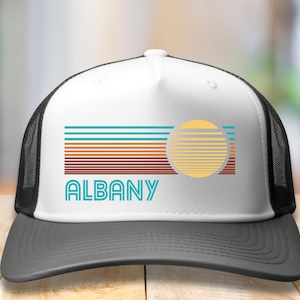 May include: White trucker hat with a black mesh back. The front of the hat features a retro-style graphic with a sun and stripes in teal, orange, and brown colors. The text "ALBANY" is printed in teal below the graphic.