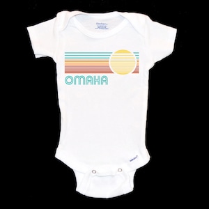 May include: White baby onesie with a retro-style graphic design featuring a sun and the word "OMAHA" in turquoise.