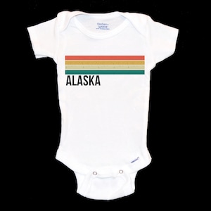 May include: White baby onesie featuring a retro-style striped design in shades of orange, yellow, and green. The word "ALASKA" is printed in black below the stripes.