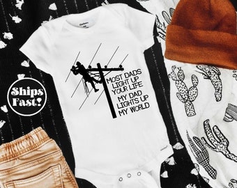 May include: White baby onesie with a black silhouette of an electrician working on a power line. The text reads "Most dads light up your life. My dad lights up my world."