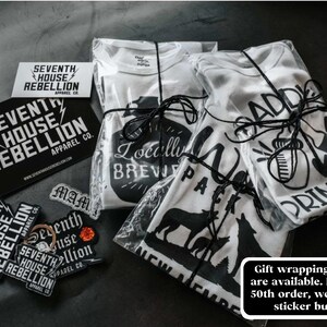 May include: Three white t-shirts with black graphic designs are wrapped in clear plastic and tied with black string. The designs include "Locally Brewed", "Wolf Pack", and "Addicted to Drinkin".  The shirts are accompanied by several black and white stickers with the text "Seventh House Rebellion Apparel Co."