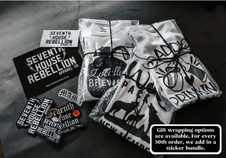 May include: Three white t-shirts with black graphic designs are wrapped in clear plastic and tied with black string. The designs include "Locally Brewed", "Wolf Pack", and "Addicted to Drinkin".  The shirts are accompanied by several black and white stickers with the text "Seventh House Rebellion Apparel Co."