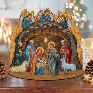 May include: A wooden nativity scene with a brown cave and a manger inside. The scene features a blue and gold-trimmed robe on Mary, a blue robe on Joseph, and a white robe on the baby Jesus. There are two angels with wings above the cave, and two sheep in the foreground.