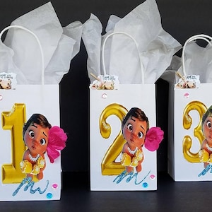 May include: Three white gift bags with handles, each decorated with a different number, 1, 2, and 3, in gold glitter. Each bag features a cartoon image of a brown-skinned girl with dark hair and brown eyes, wearing a gold necklace and a pink flower in her hair. The bags are decorated with blue and pink glitter and the text "Moana".