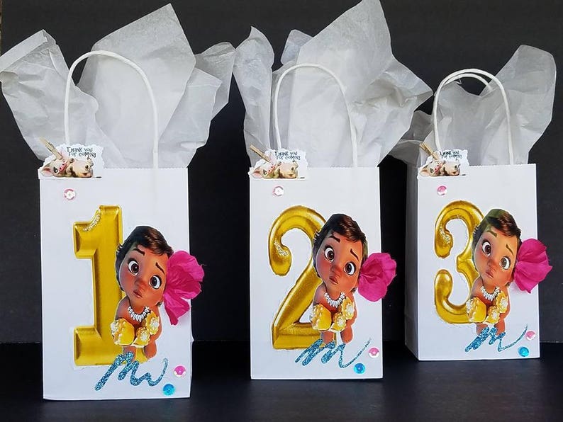 May include: Three white gift bags with handles, each decorated with a different number, 1, 2, and 3, in gold glitter. Each bag features a cartoon image of a brown-skinned girl with dark hair and brown eyes, wearing a gold necklace and a pink flower in her hair. The bags are decorated with blue and pink glitter and the text "Moana".