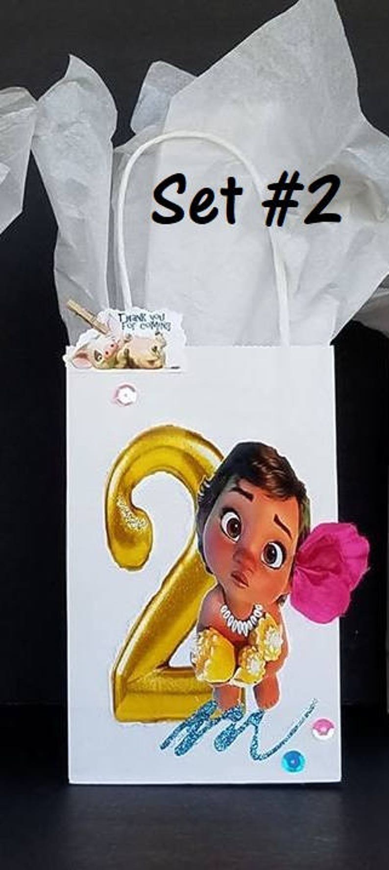 May include: A white gift bag with a gold number 2 and a cartoon character from the movie Moana. The bag has a pink flower and the text "Set #2" on the front.