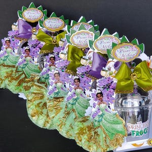 May include: A decorative centerpiece featuring a princess theme. It includes cutouts of a princess in a green dress with gold accents, purple and green bows, and the text "Princess Milan". The base has a clear jar and a frog figurine.