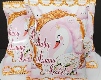 May include: Two white favor bags with a pink and gold design featuring a white swan with a gold crown, clouds, and the text "Baby Lyana Isabel".