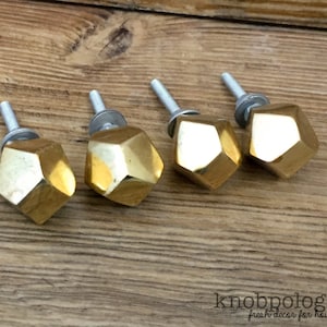 May include: Four gold-colored, faceted cabinet knobs with silver screws are arranged on a wooden surface. The knobs have a geometric, gem-like design. The knobs are for home decor.