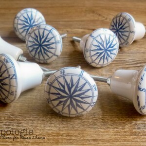 May include: White ceramic cabinet knobs with a blue compass rose design. The knobs have a cylindrical shape and are attached to metal screws. The image includes the text "knobpologie fresh decor for house & home".