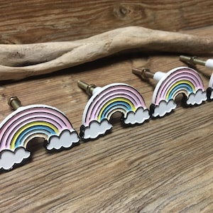 May include: Four rainbow-coloured cabinet knobs with white clouds. Each knob features a rainbow with pink, yellow, blue, and green stripes. The knobs are made of metal and have a white finish.