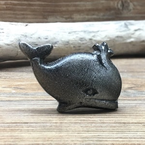 Könnte beinhalten: Ein silberner, walförmiger Schrank- oder Schubladenknopf. Der Wal ist nach rechts gerichtet und hat eine leicht strukturierte Oberfläche.