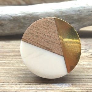 May include: A circular cabinet knob with a segmented design. It features light wood, white material, and a gold-coloured metal section. The knob is set against a wooden surface.