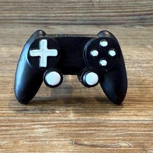 May include: A black video game controller with white buttons and a white cross-shaped directional pad. The controller has a classic design, with two handles and a series of buttons on the front. The controller is made of a smooth, matte material.