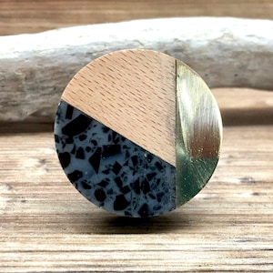 May include: A round cabinet knob with a segmented design. The knob features a light wood grain section, a section with black and gray speckled material, and a gold-colored metal section. The knob is designed for furniture.