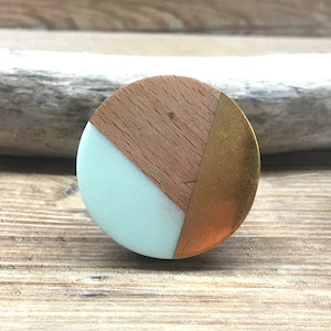 Peut inclure: Deux boutons de meuble décoratifs. Chaque bouton présente un design rond avec un dessus en bois clair, une section de résine vert menthe et une section de métal couleur cuivre. Les boutons ont une vis argentée pour la fixation et sont posés sur une surface en bois.