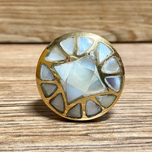 以下が含まれることがあります： 幾何学模様の丸い金色のキャビネットノブ。ノブは、三角形と台形の真珠母貝の象嵌で囲まれた中央の正方形が特徴です。ノブは木製の背景に配置されています。