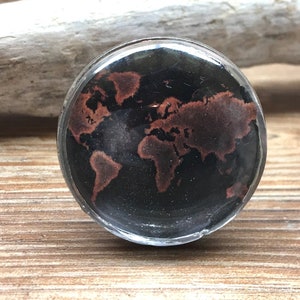 May include: A circular ring featuring a black background with a copper-coloured world map graphic. The design is encased under a smooth, clear, domed surface, set within a simple metallic band that frames the globe illustration.