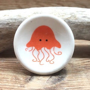 Puede incluir: Un pequeño cuenco de cerámica blanca con un diseño de medusa naranja. La medusa tiene una cabeza redondeada y tentáculos rizados. El cuenco es redondo y está sobre una superficie de madera.