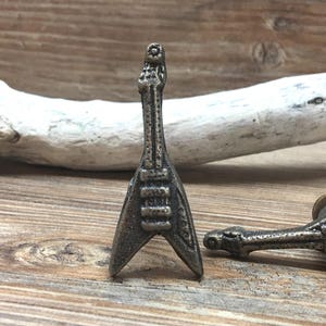 May include: Two bronze-colored guitar-shaped cabinet knobs. One knob stands upright, while the other lies on its side. The knobs are detailed with strings and other guitar features. The background is a wooden surface with a piece of driftwood.