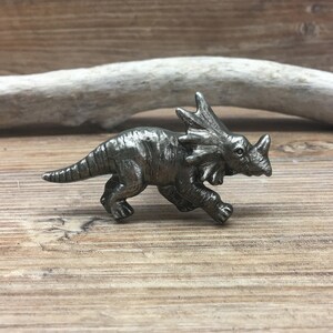 Op de afbeelding: Een zilverkleurig metalen dinosaurusbeeldje in een rennende pose. De dinosaurus heeft een kraag om zijn nek en drie hoorns op zijn kop.
