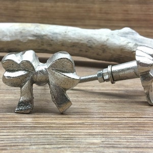 Peut inclure: Un bouton de meuble argenté en forme de nœud papillon avec un boulon fileté et un écrou. La quincaillerie décorative est en métal et est conçue pour être fixée à un tiroir ou à une porte d'armoire. Le bouton est sur une surface en bois.