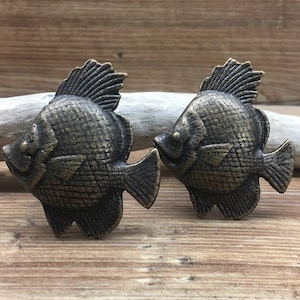 May include: Two antique gold-toned fish-shaped drawer pulls. The fish have a detailed, textured design and are facing opposite directions.