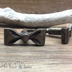 May include: Two antique bronze bow tie cabinet knobs on a wooden surface. The knobs have a detailed bow tie design and are attached to a threaded rod. The image also includes the text "Knobpologie fresh decor for house & home".