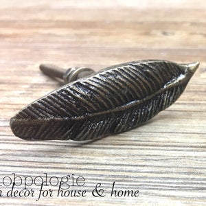 May include: Bronze-colored feather-shaped cabinet knob. The knob has detailed feather vein textures and a small, round base. The text "Knobpologie fresh decor for house & home" is at the bottom of the image.