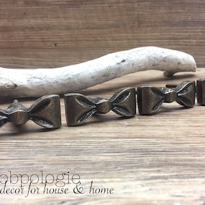 May include: Bronze-toned bow-shaped cabinet knobs arranged in a row. The knobs are made of metal and have a detailed, textured surface. The background includes a piece of driftwood and the text "Knobpologie fresh decor for house & home."